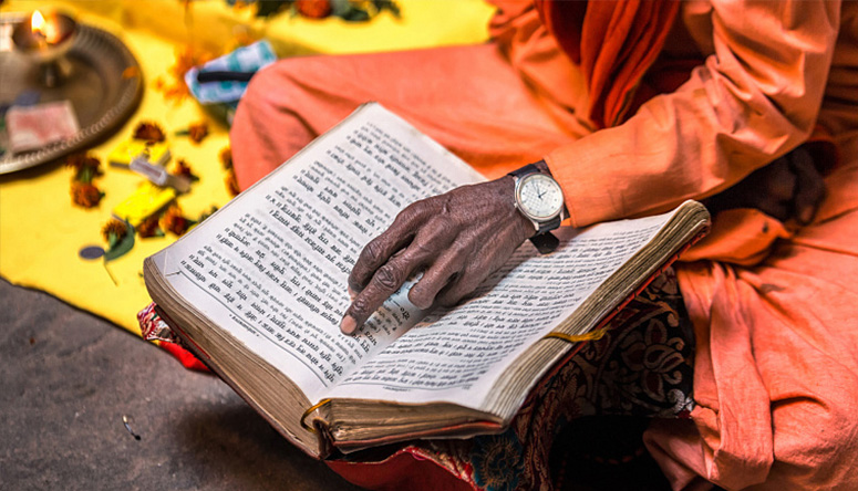 Person in orange robes reading an ancient Sanskrit manuscript to study Ayurvedic source texts and philosophy.