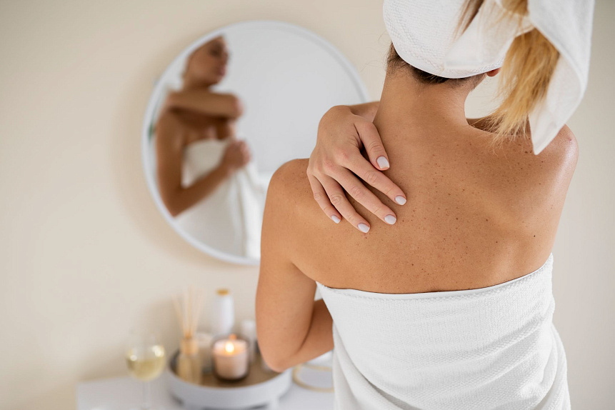 Photo of a woman from behind applying warm oil to her shoulders in front of a mirror for skin nourishment